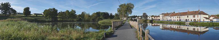 Le bief 03 pris de l'aval de la rive droite. Le réservoir de dépôt à gauche, le déversoir vers le canal sur le chemin de halage, la maison éclusière 02 et le site d'écluse 02. Deux petits lavoirs dans la rive gauche. Alignements de maisons le long du chemin de halage devenu route départementale.