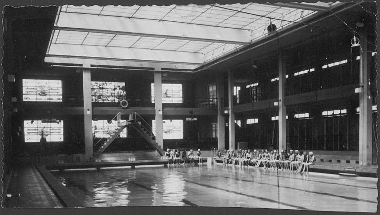 Vue intérieure de la piscine pasteur de Maubeuge avant un cours de natation. Dans le fond, noter le plongeoir et les vitraux représentant des scènes marines. Photographie, vers 1940 (AC Maubeuge).