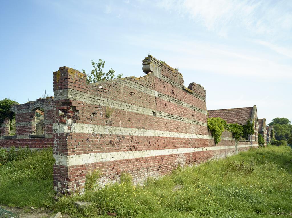 Mur de cloture sud de la ferme avec des pignons des bergeries et porcheries en prolongement.