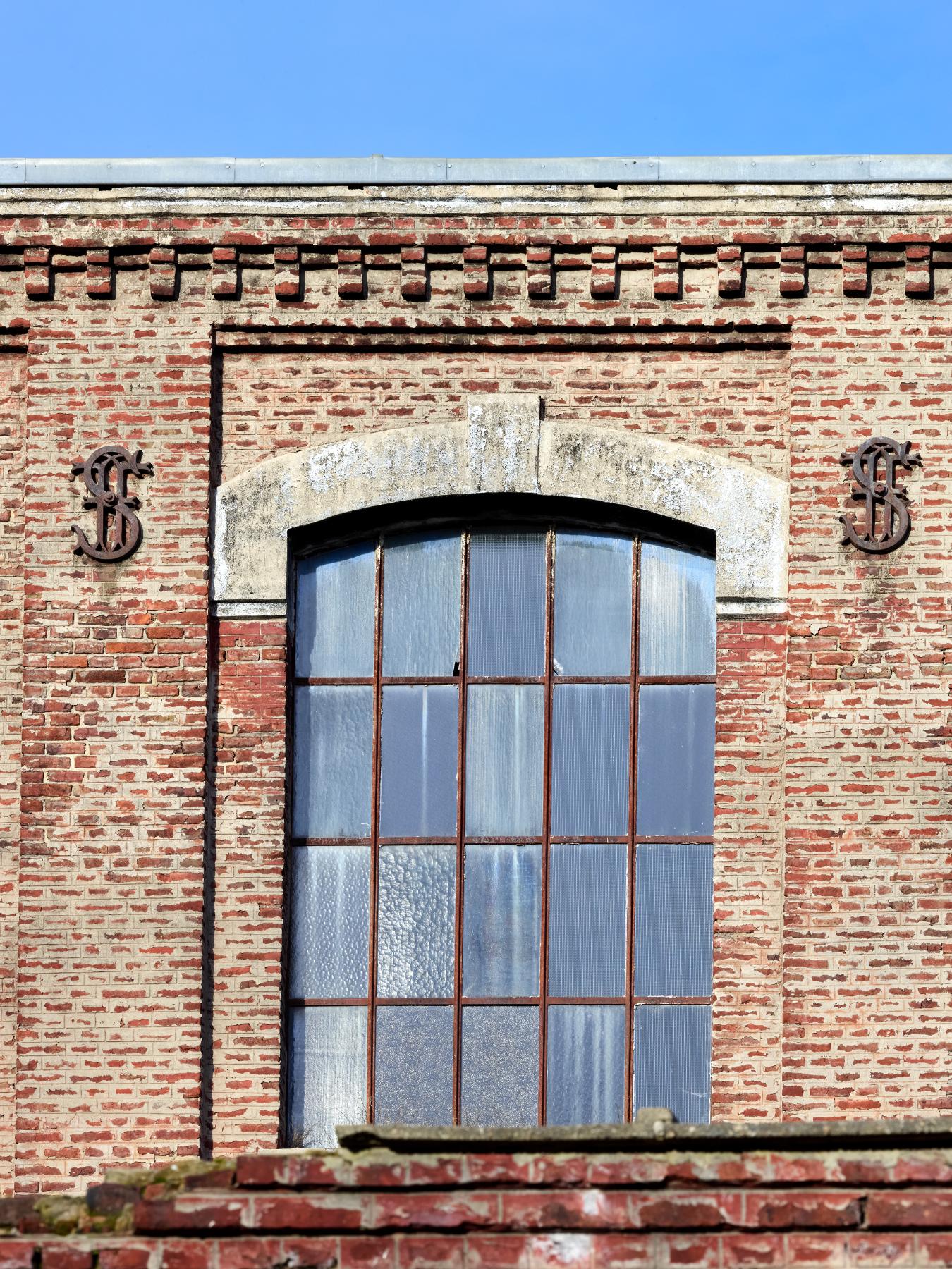 Détail d'une ouverture du deuxième étage de l'ancien atelier de fabrication encadré de fers d'ancrage portant les initiales de l'industriel. 