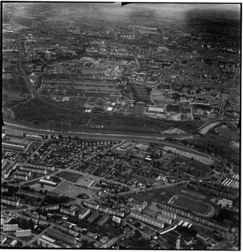 Vue aérienne : la ville vue du nord-est. Bourtzwiller au premier plan, l'usine Manurhin à droite, de l'autre côté la Doller, le cimetière central et la Cité Jean Wagner au centre.