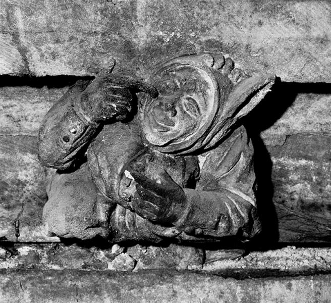 Façade sud, relief sur la corniche du bas côté, caché par la chapelle de la Vierge : fou coiffé d'un bonnet montrant son nez de son index droit (S6)