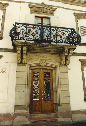 Vue de détail de la porte d'entrée avec linteau surmontée d'un balcon soutenu par 2 paires de consoles ouvragées.