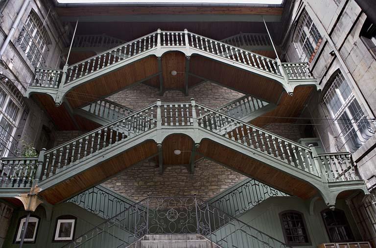 Escalier à cage ouverte à droite de la première cour : vue d'ensemble, de face, des volées suspendues en charpente.
