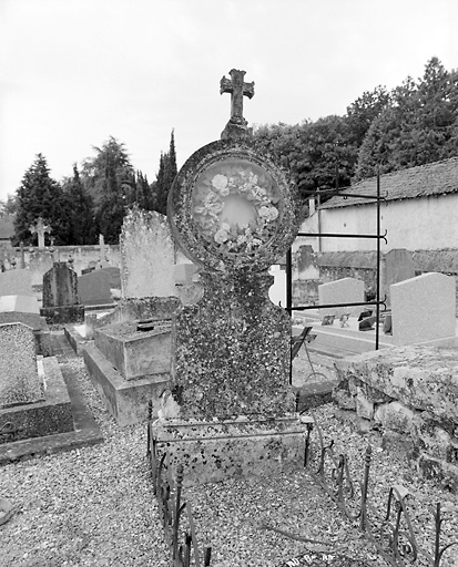 Inhumation en 1887. Stèle surmontée d'un large médaillon circulaire contenant une couronne artificielle protégée par du verre.