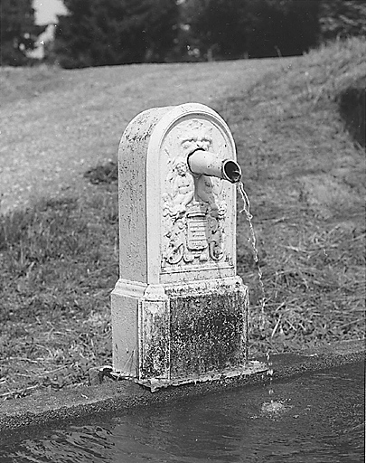 Le Puy-Merlin. Fontaine, abreuvoir, lavoir. Vue de détail de la fontaine en fonte, ornée d'une tête de lion et d'un blason, cantonnés de deux personnages mythologiques. Inscription, en partie illisible, mentionnée sur la fontaine : ACHILE..../ PARIS.