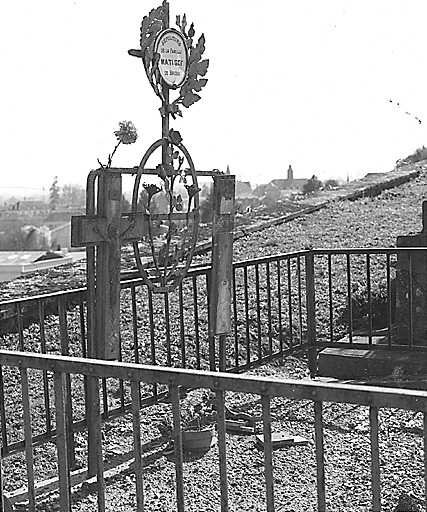 Tombe, section I. Vue de deux décors en fer forgé, superposés, exécutés vraisemblablement à la fin du 19e siècle ou au début du 20e siècle ; l'un représente des feuilles de laurier qui encadrent une plaque funéraire fixée à une croix et l'autre, un décor végétal fixé à une armature ovale.