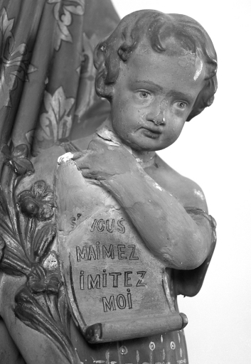 Groupe relié en plâtre polychrome représentant saint Joseph et l'enfant Jésus. Vue de détail du parchemin tenu par l'enfant Jésus, portant l' inscription peinte : 'SI VOUS / MAIMEZ / IMITEZ / MOI '.