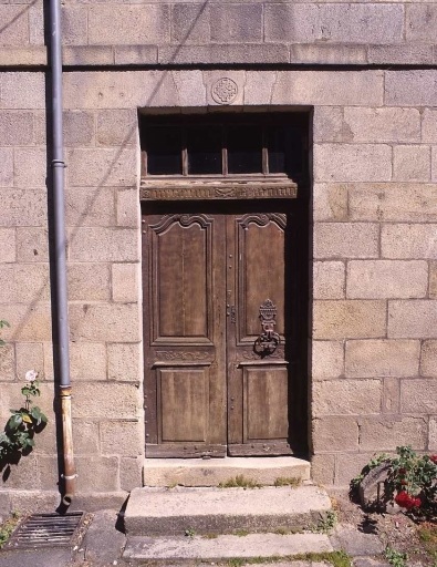 Vue de détail de l'élévation antérieure montrant la porte d'entrée. Cette porte aux moulurations chantournées présente un décor floral sur la traverse des vantaux et de l'imposte ; le vantail droit a conservé son heurtoir orné d'un monogramme.