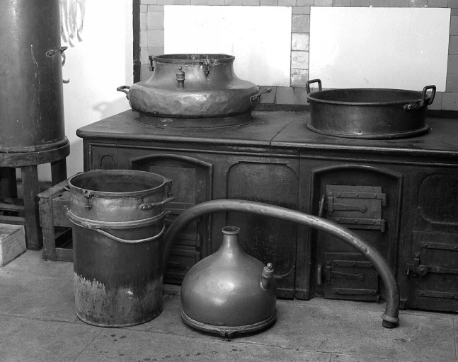 Un alambic démonté : au sol, de gauche à droite, le bain-marie, le chapiteau et le col de cygne ; en place dans le fourneau, la curcubite (le récipient à droite est une bassine à sirop) ; à gauche du fourneau, le réfrigérant.