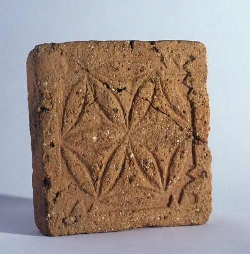 Carreau en terre cuite récupéré au moment de l'aménagement du jardin créé au sud de l'église, sur l'emplacement de l'ancien cloître. Vue de la face ornée d'un motif gravé représentant six lobes inscrits dans un cercle.