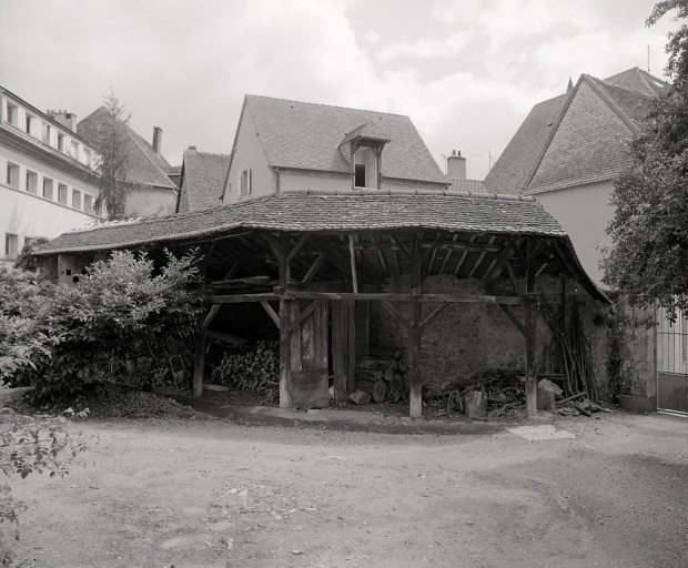 Vue d'un hangar (peut-être remise à chevaux), adossé au mur de clôture de la cour et dont la couverture en tuile creuses repose sur des poteaux en bois.