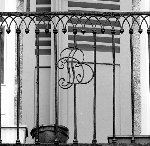 Elévation antérieure donnant sur la place. Vue de détail du monogramme D. B. ornant le garde-corps en fer forgé du balcon du premier étage.