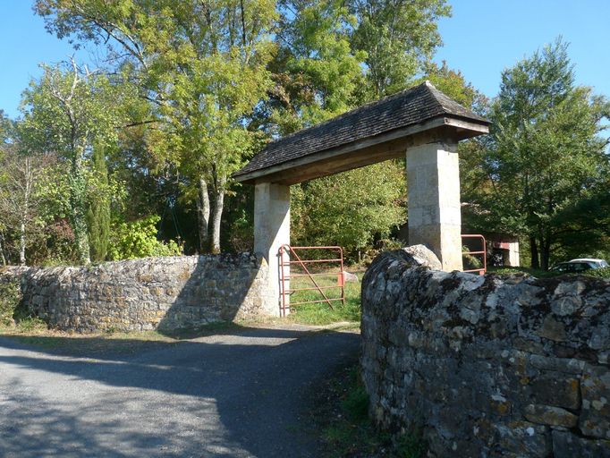 Mur de clôture à chaperon triangulaire et portail couvert d'un toit à croupes encadré de deux piliers en pierre de section carrée.