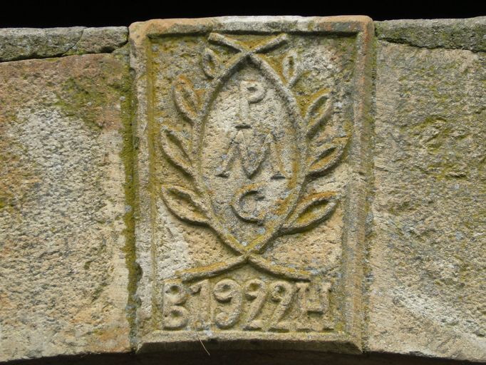 Détail de la clef du linteau de la porte charretière portant la date 1922  encadrée des initiales B.H et surmontée des lettres P.M.C entourées de rinceaux de lauriers. ; La Martinie. Clef sculptée portant la date 1922  encadrée des initiales B.H et surmontée des lettres P.M.C entourées de rinceaux de lauriers.