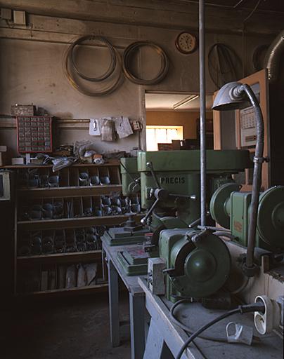 Intérieur de l'atelier : 2 machines à percer au premier plan, les fournitures au second plan avec la porte qui s'ouvre sur le bureau.