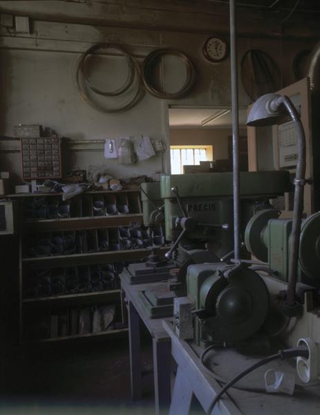 Intérieur de l'atelier : 2 machines à percer au premier plan, les fournitures au second plan avec la porte qui s'ouvre sur le bureau.