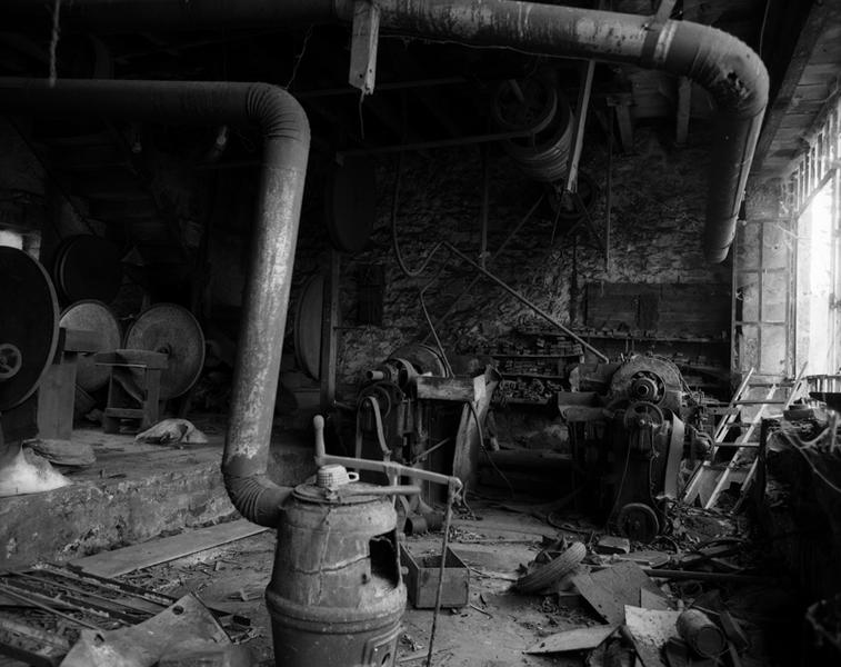 Vue intérieure de l'atelier : poêle de chauffage au premier plan, 2 machines à émoudre au second plan, axe de transmission en hauteur.