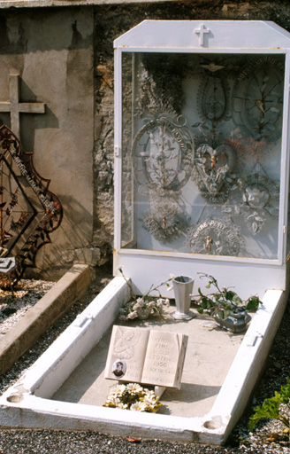 Le cimetière. Une tombe d'enfant datée 1956. Nombreuses couronnes de perles protégées par une vitrine dont les montants sont peints en blanc.