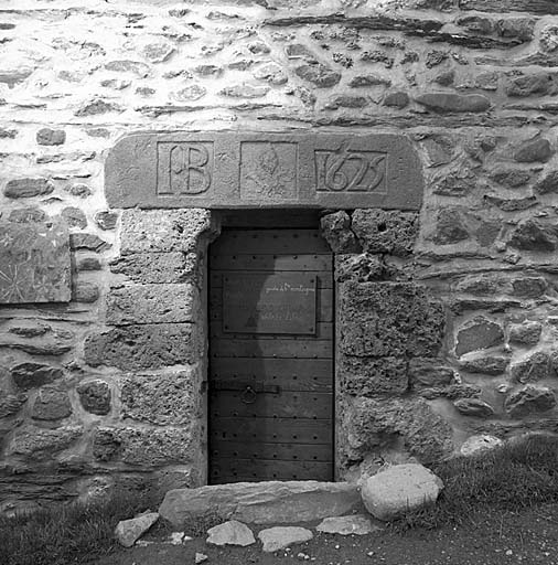 Porte d'une anciene chambre du bourg de La Grave (parcelle 396). Sur les chambres datées du XVIIe siècle, les linteaux sur coussinets reposent sur des piédroits construits en tuf sans alternance avec des plaquettes d'ardoise. La fleur de lys, au centre de l'inscription, a été bûchée.