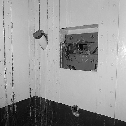 Bloc 1 (entrée). Caponnière de défense d'entrée, vue intérieure. Créneau de fusil mitrailleur R.B. A gauche, le tube (sans tête) de goulotte à grenades. En bas, le tube dévacutation des étuis du F.M. Au mur, un revêtement métallique.