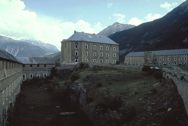 Intérieur du fort vu de l'angle sud-ouest. A gauche, la façade de la caserne O, au centre, le bâtiment E (ancienne chapelle), à droite, la caserne B, à l'arrière-plan, au centre, le piton du Janus et, à droite, les sommet et fort de l'Infernet.