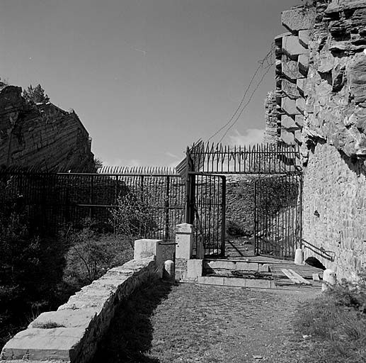 Fort supérieur. Front de tête. Porte. Ensemble vu de l'extérieur, depuis la route d'accès. Au premier plan, le haha et le pont roulant en partie effacé dans sa casemate. Derrière la grille défensive et l'avant-porte. A droite, corbeaux-supports du pas de souris de jonction avec la batterie des Caurres. A l'arrière-plan, à gauche, rocher de l'escarpe et double caponnière.