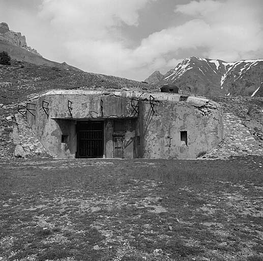 Bloc 1 (entrée). Vue d'ensemble. De gauche à droite : créneau FM de caponnière ; entrée du matériel (pont-levis relevé), entrée du personnel, dans l'ombre, créneau JM, puis cheminée des gaz brûlés. Au-dessus, cloche GFM. Dans la visière, supports de l'antenne radio.