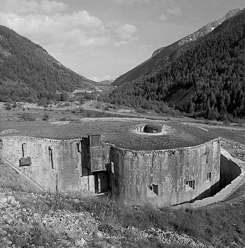 Vue d'ensemble de la gorge. Caponnière de gorge avec : à gauche, l'entrée de l'ouvrage et la cheminée d'accès d'hiver ; au-dessus, la cloche GFM arrière ; au-dessus des créneaux FM de droite, prise d'air de la ventilation.