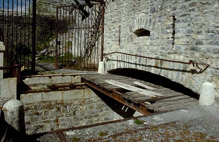 Fort supérieur. Front de tête. Porte. Vue rapprochée prise de l'extérieur. Au premier plan, le haha coupant la route et le pont roulant en partie effacé dans sa casemate. Derrière, la grille défensive et l'avant-porte. A l'arrière-plan, la double caponnière.