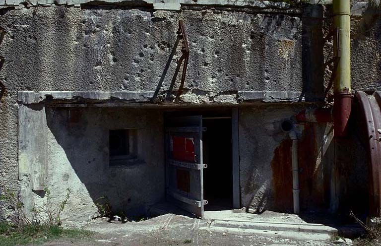 Bloc 1. Vue de face. Façade avec la porte d'entrée et l'arrivée de la voie de 0,60 venant du téléphérique. A gauche, caponnière et créneau de FM de défense rapprochée. Au-dessus, visière et supports de l'antenne radio.