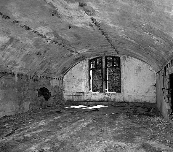 Vue intérieure d'une casemate logement du premier étage prise depuis le couloir de fond en direction de la façade. En plafond, crochets de fixation des tôles-parapluies.