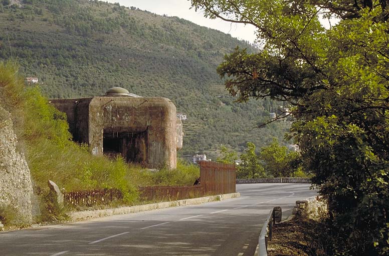 Bloc actif (B 4). Flanc droit, vue extérieure. Créneau du JM 5 sous casemate. A l'arrière plan, mont et ouvrage de l'Agaisen. Au premier plan, la route D 2204 et la grille défensive du bloc. ; Sospel, ouvrage mixte de Saint-Roch.