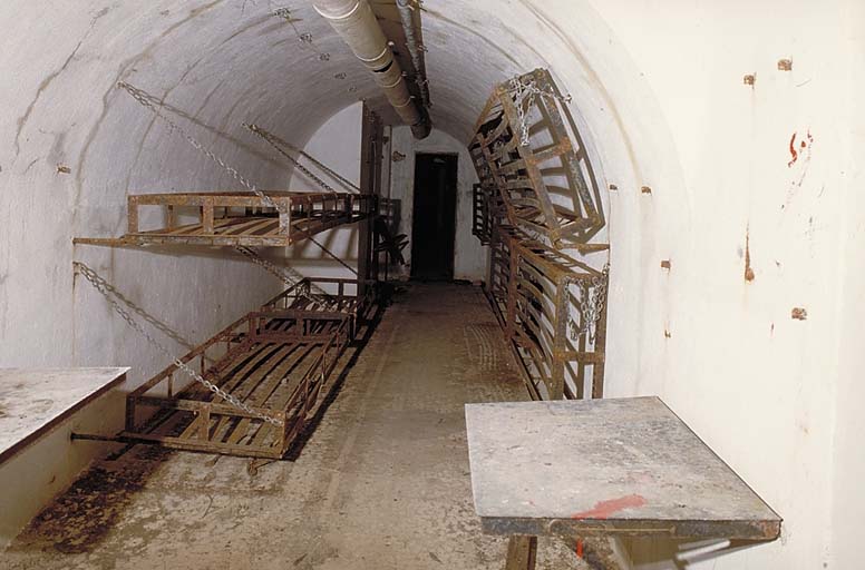 Galerie centrale, vue intérieure. Au mur, couchettes à rabattement. Au plafond, gaine de ventilation et collecteur des fumées des lanternes d'éclairage. Au fond, porte d'accès à la casemate double de mitrailleuse. ; Sospel, ouvrage de Castes Ruines. Vue intérieure de la galerie centrale.