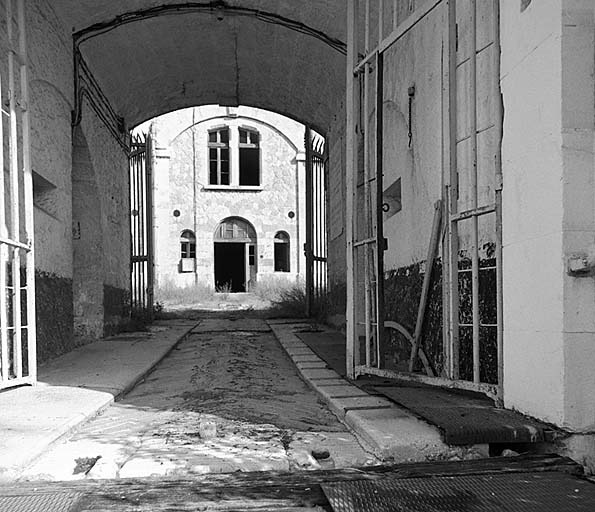 Passage d'entrée. Vue d'enfilade depuis l'extérieur avec les deux portes-grilles successives. Au fond, cour centrale et caserne d