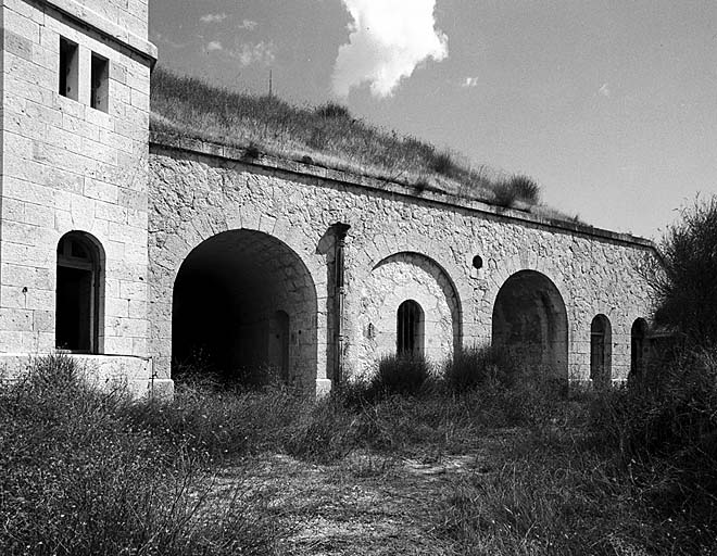 Bâtiment m. Vue d'ensemble de la façade. A gauche, extrémité de la façade de la caserne d, puis passage couvert menant au front de tête. Au 1/3 à droite, arcature formant visière de la fenêtre d'éclairage du magasin à poudre