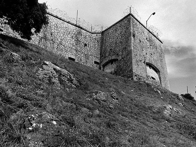 Aile gauche du bâtiment a. Vue extérieure prise en contrebas. Face gauche (nord-ouest) : créneau de pied surmontant une embrasure à canon flanquant l'escarpe du front sud-ouest. Face droite (sud-ouest) : créneau de pied surmontant une embrasure à canon tirant sur la région du col d'Eze.