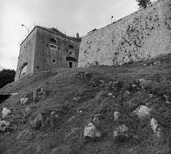 Extrémité gauche du bâtiment a. Vue extérieure prise en contrebas de la route d'accès. De gauche à droite : créneau de pied surmontant une embrasure à canon tirant sur le col d'Eze (face gauche). Face droite : en bas : embrasure à canon. Créneau de pied surmontant la poterne encadrée de deux créneaux de fusillade verticaux. En haut : deux groupes de créneaux de fusillade (un créneau horizontal entre deux créneaux verticaux) assurant la défense rapprochée.