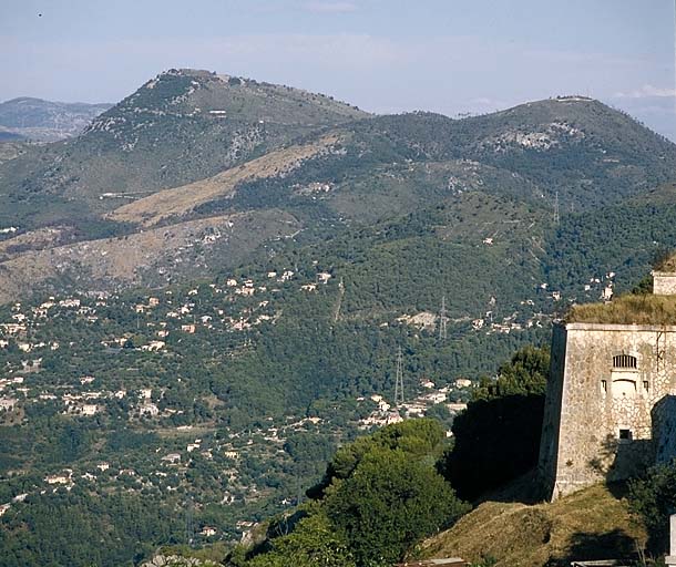 Vue lointaine des monts chauves prise des abords du fort. Au premier plan, à droite, extrémité du bâtiment a. Au deuxième plan, à gauche, fort du mont chauve d'Aspremont. A droite, ouvrage du mont chauve de Tourette