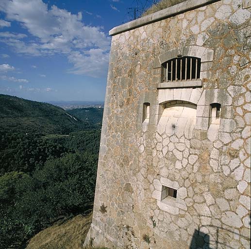 Extrémité gauche du bâtiment a vue de l'extérieur. En bas, embrasure à canon battant la pente en contrebas de la plateforme d'entrée. Au-dessus, ensemble de créneaux de fusillade (un horizontal et deux verticaux) de défense rapprochée de l'entrée.