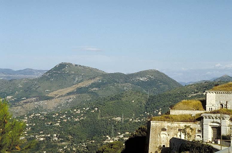 Vue lointaine des Monts Chauves prise des abords du fort. Au premier plan, à droite, extrémité du bâtiment a. Au deuxième plan, à gauche, fort du Mont Chauve d'Aspremont. A droite, ouvrage du Mont Chauve de Tourette.