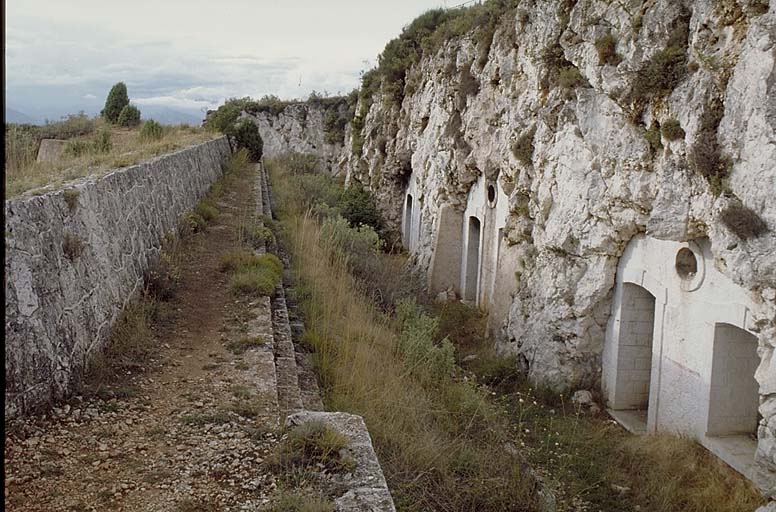 Façades des abris cavernes et cour centrale. A gauche, banquette et parapet d'infanterie du front de gorge. ; Tourrette-Levens, ouvrage du Mont Chauve : façades des abris-cavernes.