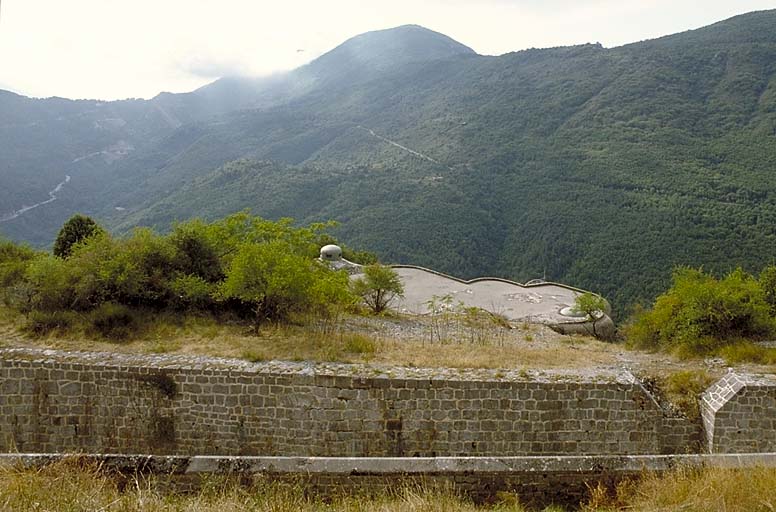 Vue prise du parapet du front sud. Au premier plan, mur à bahut du parapet d'infanterie et fossé, avec en avant le bloc 2 de l'ouvrage moderne. Au fond, col et ouvrage de Castillon avec, à gauche, la pointe de la Pena et à droite le mont Ours.