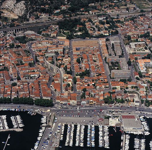 Partie ouest. Vue aérienne prise du sud en direction du nord. Au premier plan le port et la section du chemin du Littoral dite Plage de l'Estaque (RN 568b), à l'arrière-plan la voie ferrée. Le quartier est délimité à gauche par le boulevard Roger-Chieusse qui sépare la Fontaine-des-Tuiles du quartier de l'église. A droite, la résidence Saumaty, construite en 1974 à l'emplacement de la tuilerie Pierre.