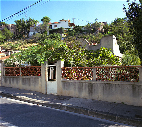 Vue de la clôture, du portail, du jardin et du fronton de la maison émergeant de la végétation.