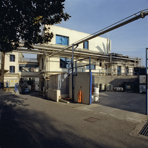 Vue depuis la rue intérieure :  les ateliers surélevés dans les années 1990 (15 et 13) et la passerelle de communication. A droite, l'ancien bassin des eaux de rétention. Voir Doc. 06.