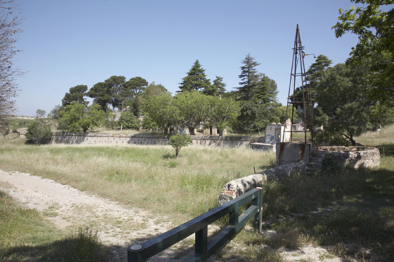 Vue d'ensemble prise du sud. Au premier plan : puits et éolienne ; au second plan : terrasse en terre-plein avec maison du jardinier, orangerie et annexes ; à l'arrière-plan : les arbres devant les bâtiments principaux.