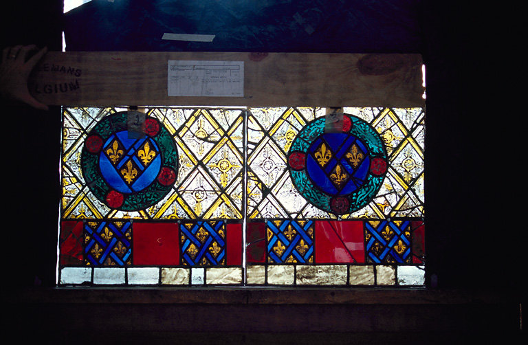 verrière : Panneaux 2, 3 (Ap. R.) La bordure inférieure est une alternance de motifs bleus et rouges entre deux filets blancs. Vers le centre du vitrail, un motif de losanges orné de feuilles quadripétales. Sur ce motif est placé une couronne de feuillages dont le fond, bleu, est orné de trois fleurs de lys.