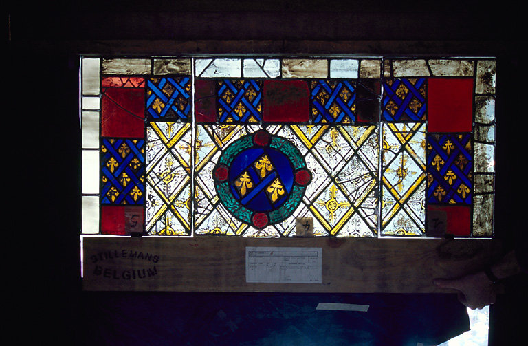 verrière : Panneaux 1, 4, 5 (Ap. R.) angles de la bordure, alternance de motif bleus et rouges entre deux filets blanc ; grisaille à treillage losangé, une couronne de feuillages entoure trois fleurs de lys sur un fond bleu.