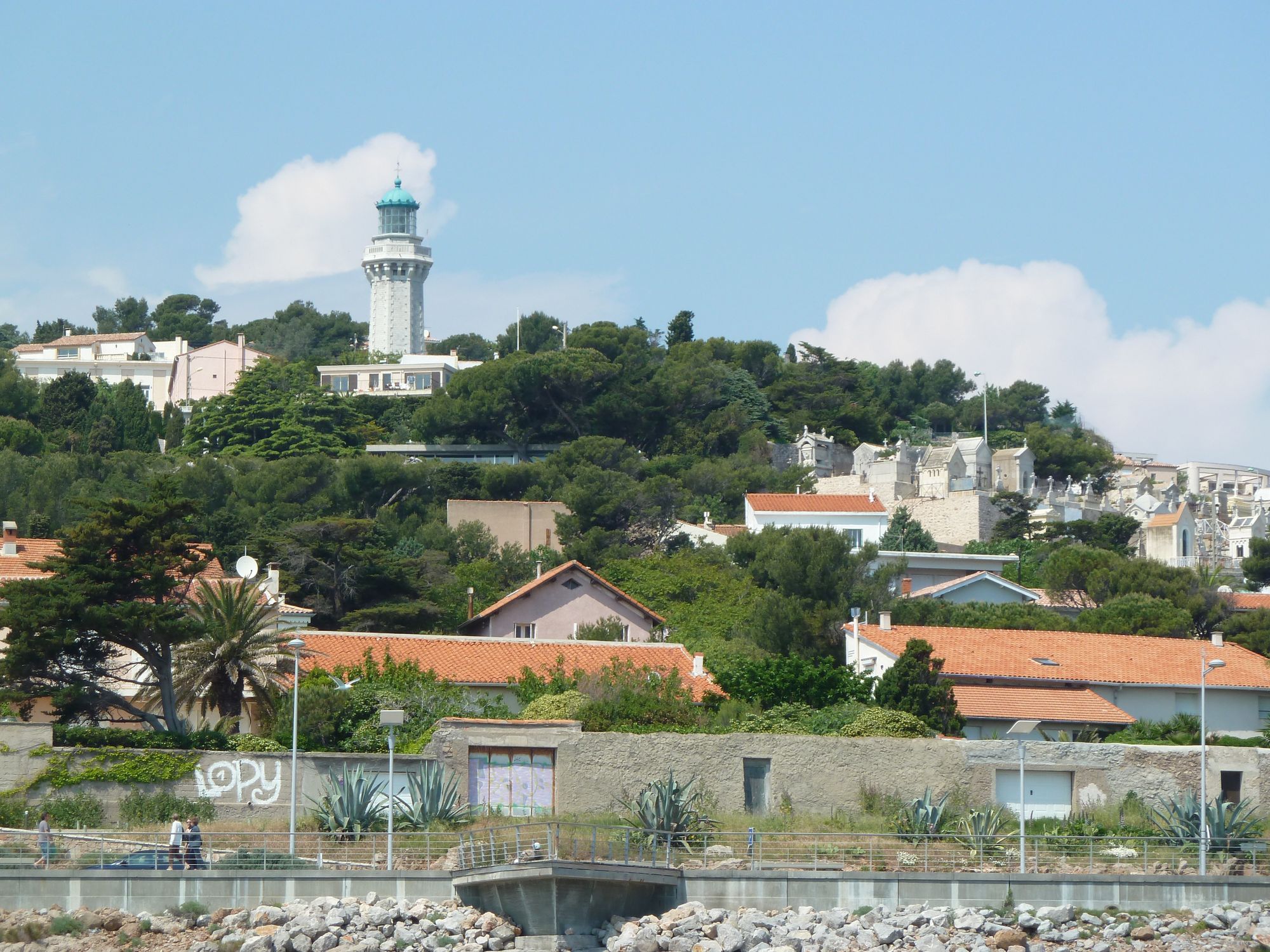 Vue de la villa depuis la mer, cachée par la végétation, un peu en dessous du phare Saint-Clair. On aperçoit uniquement le bandeau horizontal de son toit terrasse.